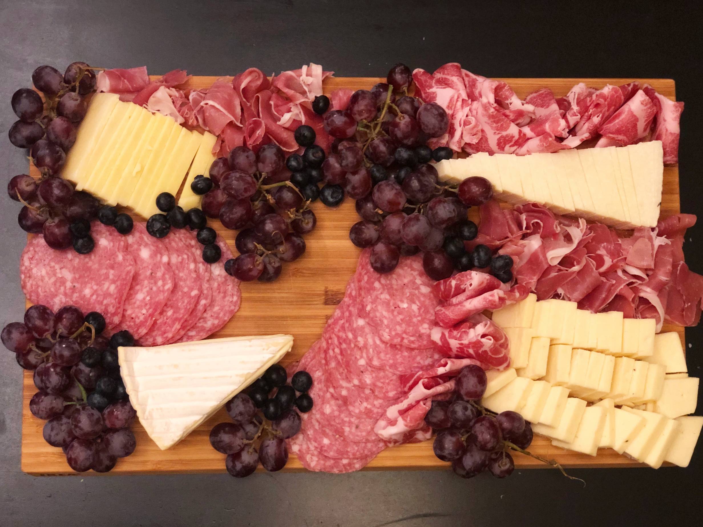 A wooden cutting board with meats, cheeses, and added fruits. Grapes in small clusters and small groups of blueberries are touching the cheese and meats. Photo by Alyssa Buckley.
