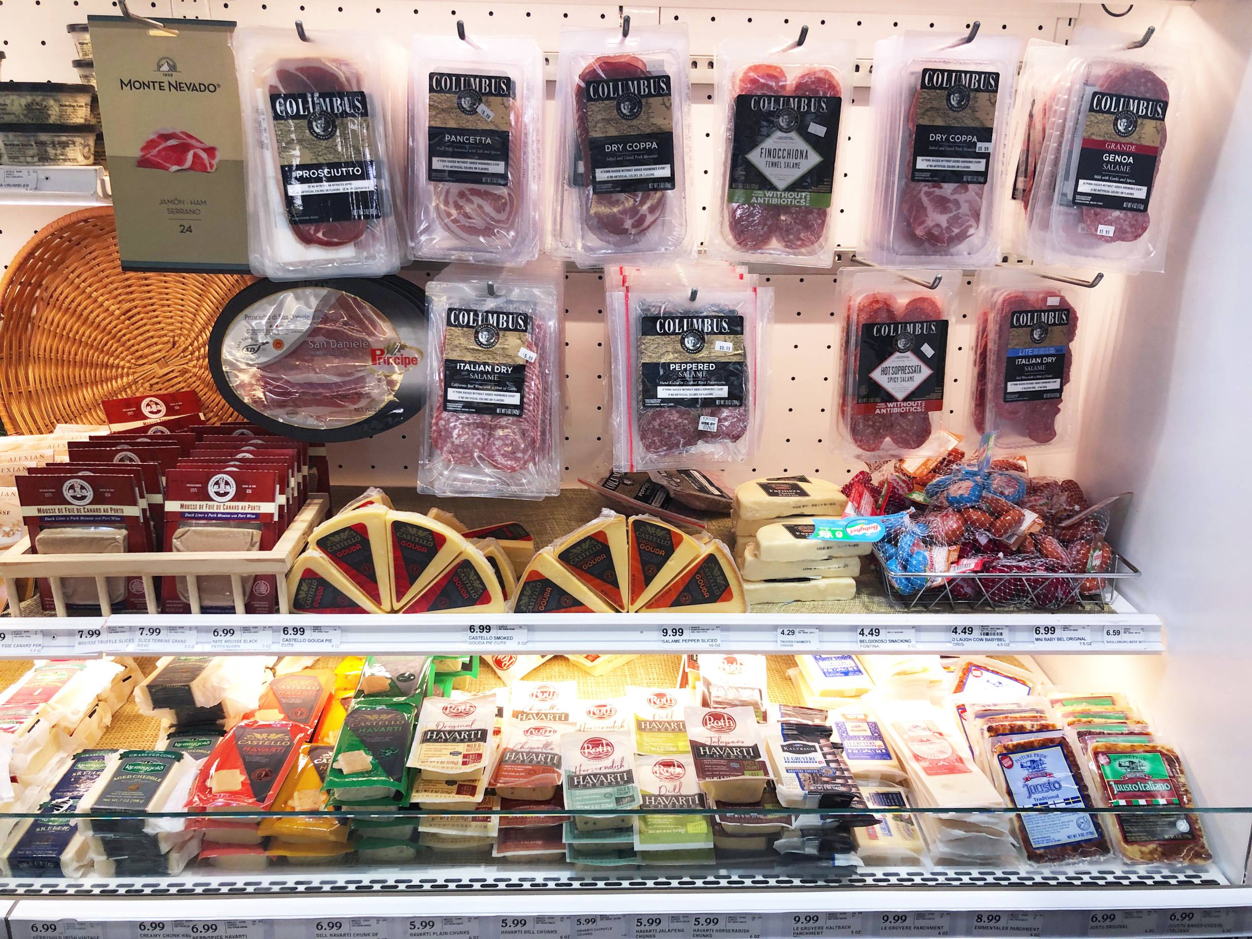 The meat section of Harvest Market has many packaged deli meats hanging from metal hooks. Below the meat are more wedges and rectangles of packaged cheeses in a white display case. Photo by Alyssa Buckley.