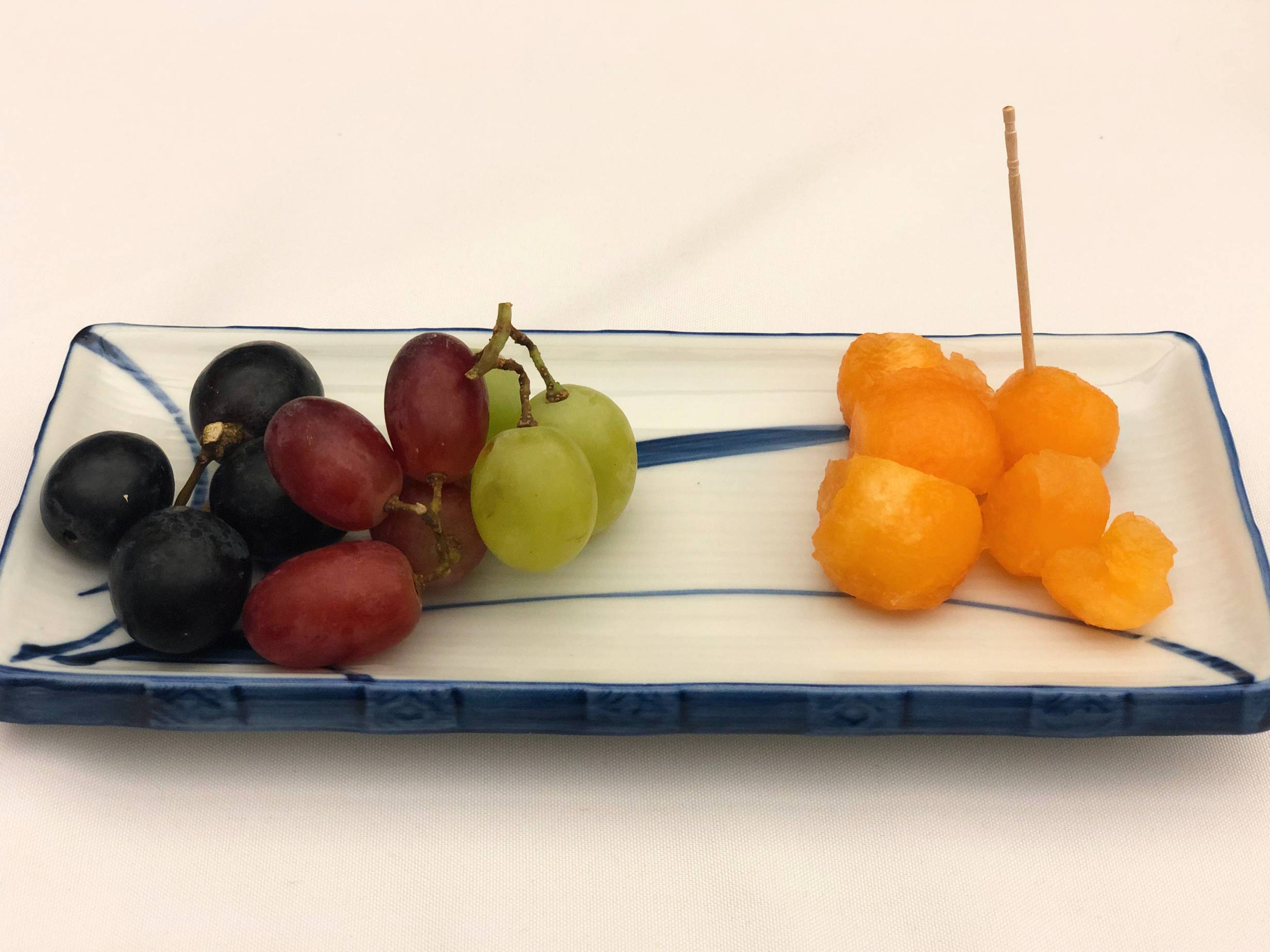 Three black grapes, three red grapes, and three green grapes on the vine are clustered on the left side of a rectangular ceramic plate. On the left, there are balls of cantaloupe with one toothpick stuck in a ball. Photo by Alyssa Buckley.