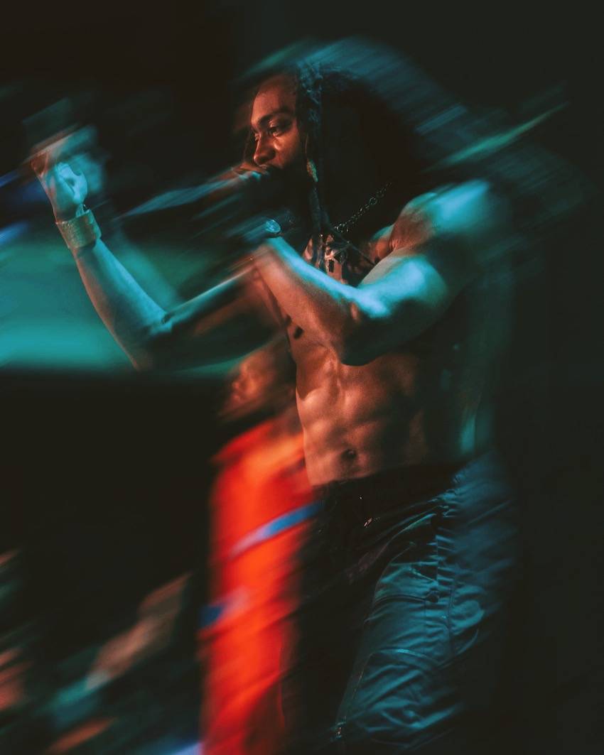 A long exposure photo that emphasizes the teal and red stage lights. The motion of the rapper gesturing with his hand is captured in the long exposure.