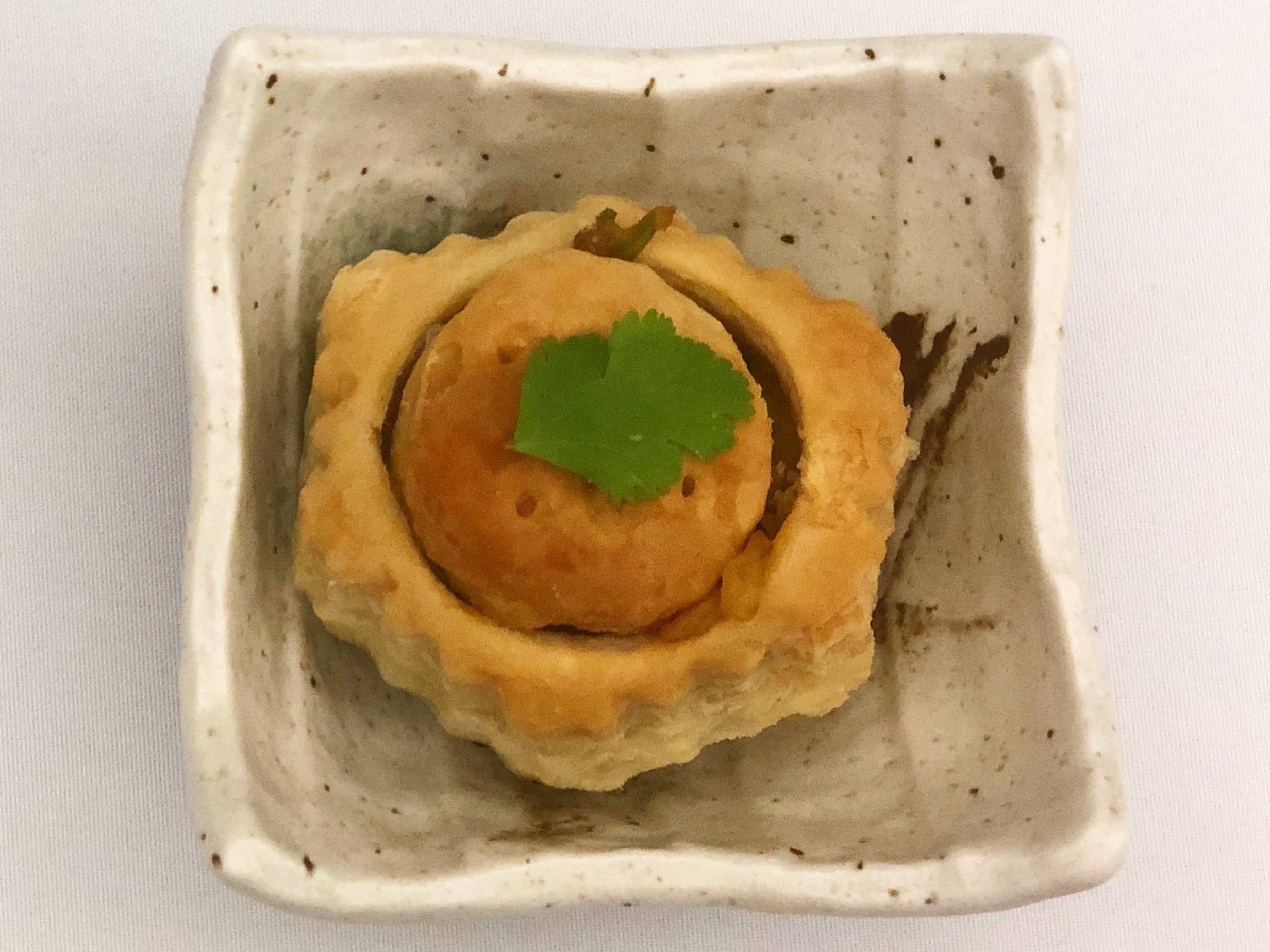 A vegetable puff pastry sits in a square plate. The puff is divided in the center as a circle with a small leaf on top. The plate is on a table with a white tablecloth. Photo by Alyssa Buckley.