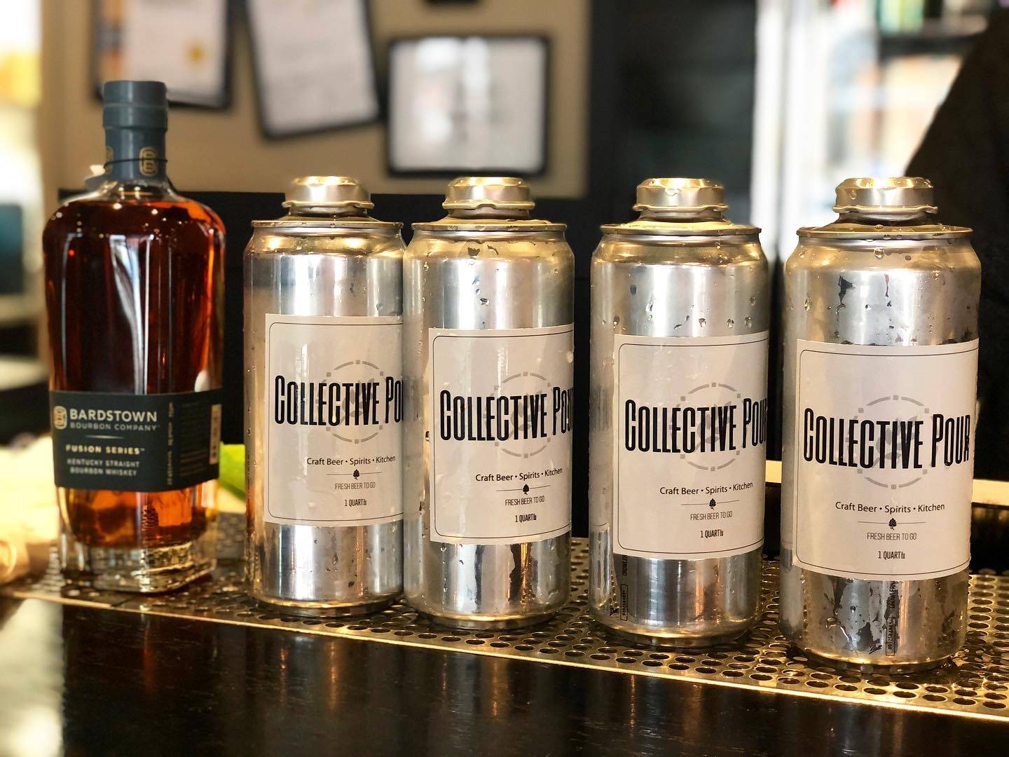 A bottle of brown whiskey sits next to four silver crowler cans with a sticker label reading 