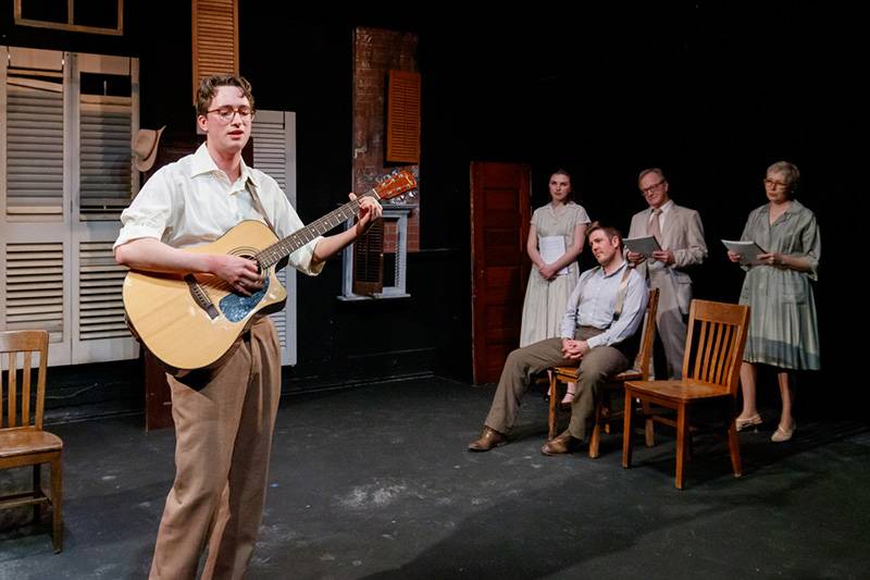  Photo of actor singing and playing guitar to the left with four actors seated and listening off to the right. Photo by Jesse Folks.