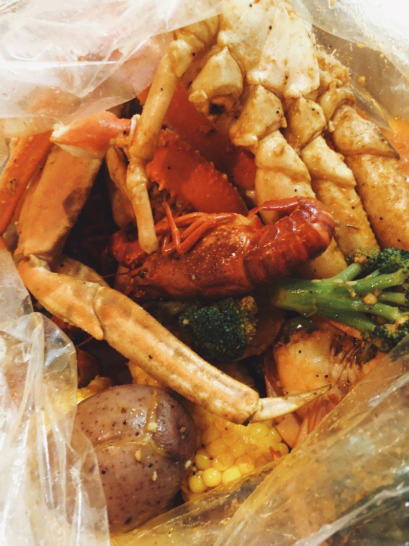 A plastic bag is filled with crab legs, shrimp, red potatoes, corn on the cob, and broccoli in a thin sauce. Photo by Remington Rock.
