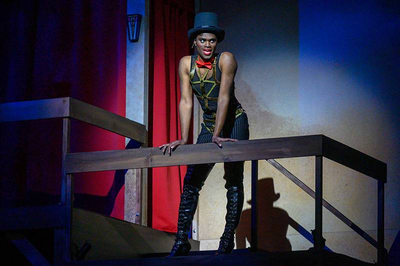 Image: Photo from Cabaret with Corey Barlow as Emcee in black leotard, tights, boots, and top hat leaning over the balcony. Photo by Darrell Hoemann. 