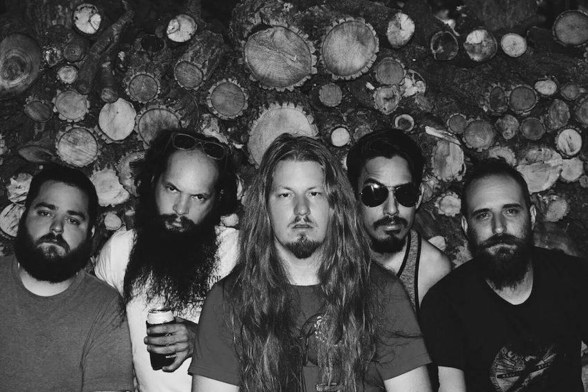 A black and white picture of the five members of Carrie Nation and the Speakeasy. All the members sport either a beard or a goat-tee. The stare blankly into the camera. The member second from the left holds a beer in a koozie, the member second from the right wears aviator sunglasses with one eyebrow cocked.