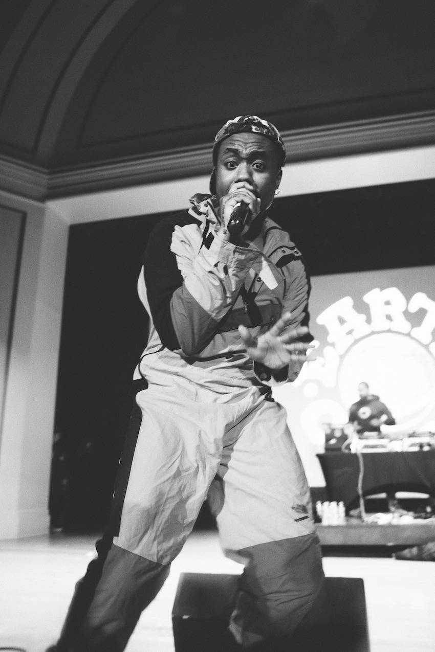 A member of EarthGang wears an Eighties-style track suit. He raps into a microphone and looks directly into the camera. The DJ for the group can be seen in the background. The Photo is black and white.