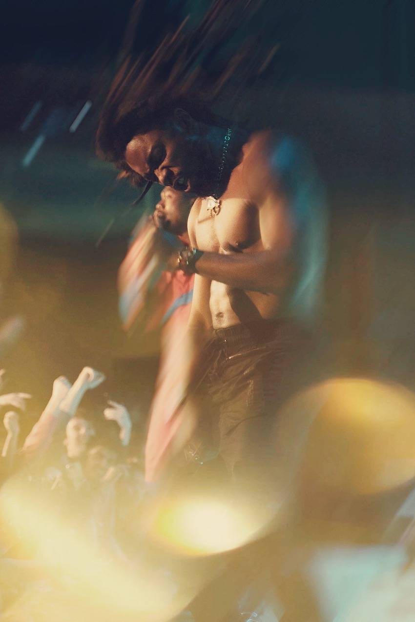 A long exposure photo of a shirtless rapper making a fierce face as if he is grunting. The movement of his dreadlocks is caught in the stage lights. 