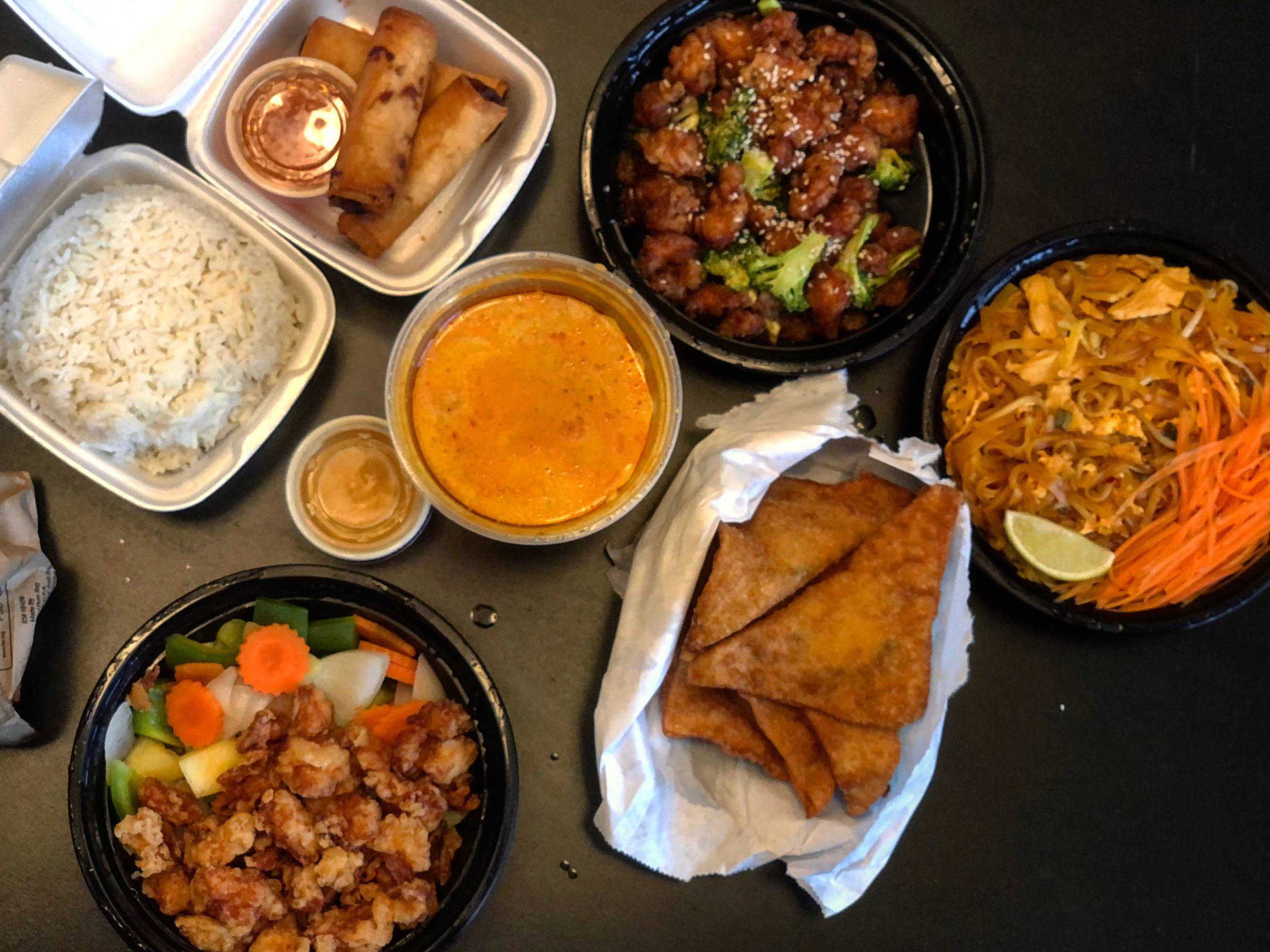 A variety of takeout dishes lay on a black table. A container of steamed white rice in a styrofoam square container, a small orange sauce in a plastic cup, a yellow curry in a plastic container, and two chicken dishes in black plastic circle containers. There is pad thai in a black circuluar plastic container, three egg rolls in a styrofoam container, and several triangular crab rangoons. Photo by Alyssa Buckley.