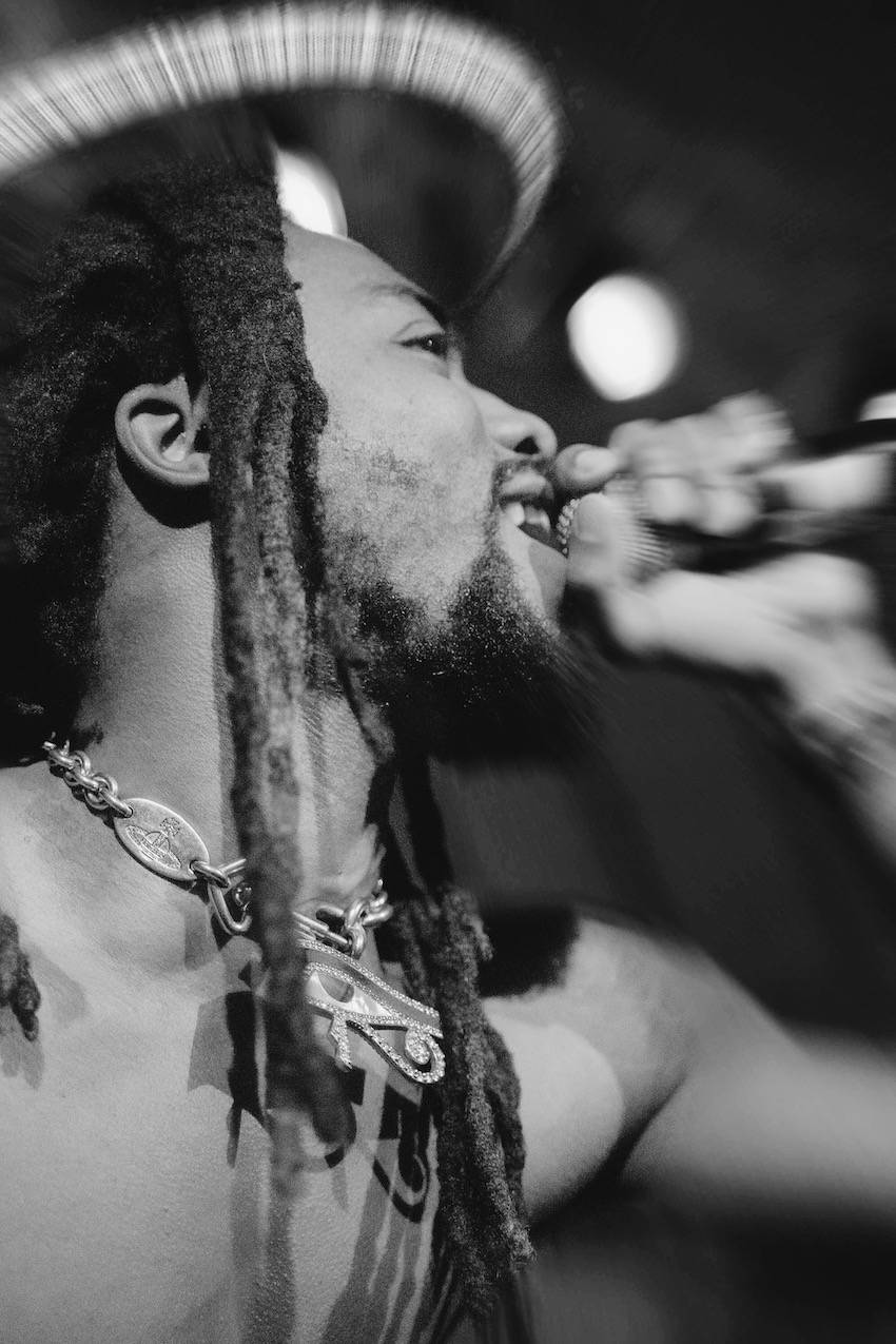 A close-up on one of the rappers, shirtless. An Egyptian-eye necklace hangs around their neck. The edges of the photo are blurry, obscuring his hand with the microphone and his hat.