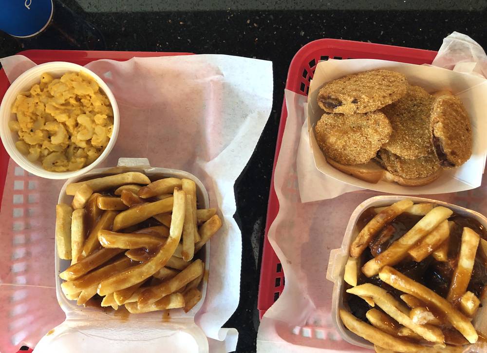 At Wood N Hog Barbecue in Champaign, two red plastic trays with white paper lining each hold a square Styrofoam container of meat and sauce topped with French fries. One tray has a side of macaroni and cheese; the other tray has a side of fried green tomatoes. Photo by Jessica Hammie. 