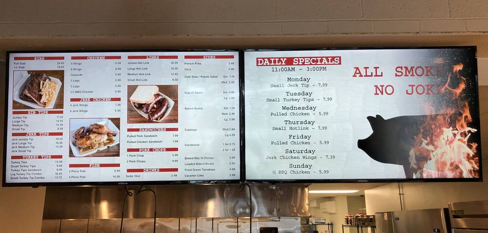 Digital menu boards at Wood N Hog Barbecue. On the left side is a list of menu items and prices. On the right side is a list of daily lunch specials and prices. The menus hang over the register, in front of the open kitchen. Photo by Jessica Hammie. 