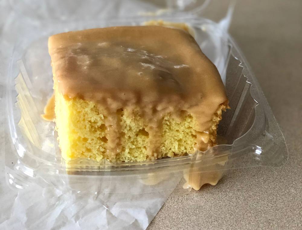 Caramel cake at Wood N Hog is served in a plastic to-go container. A square piece of yellow cake is topped with a light brown caramel glaze. Some of it drips down the side. Photo by Jessica Hammie. 