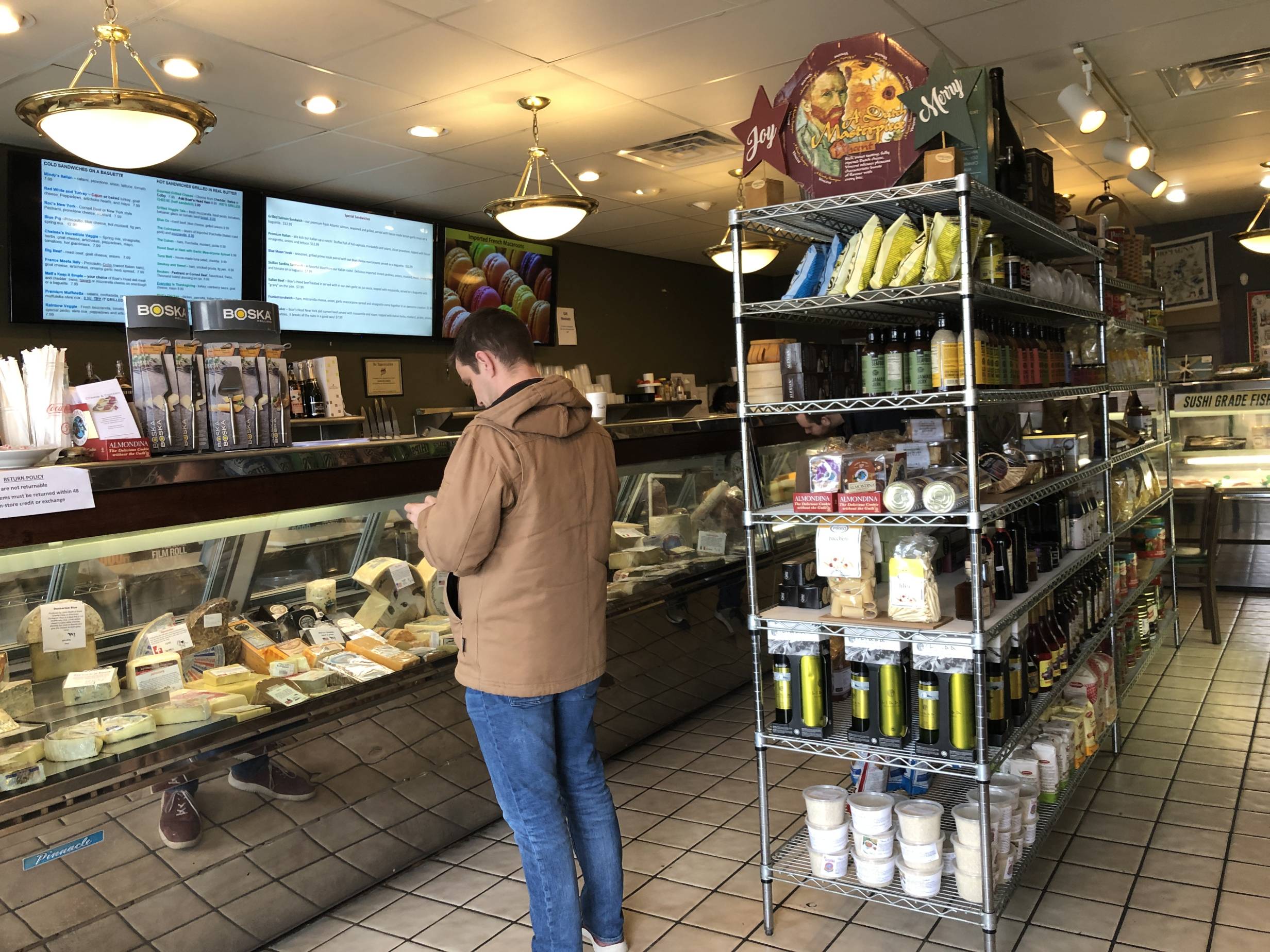 The interior of Cheese & Crackers with a patron waiting to be served. There is a long refrigerated case of various cheeses followed by another of cured meats and sausages. The middle has a many-tiered shelf of non-perishable goods. Photo by Alyssa Buckley.