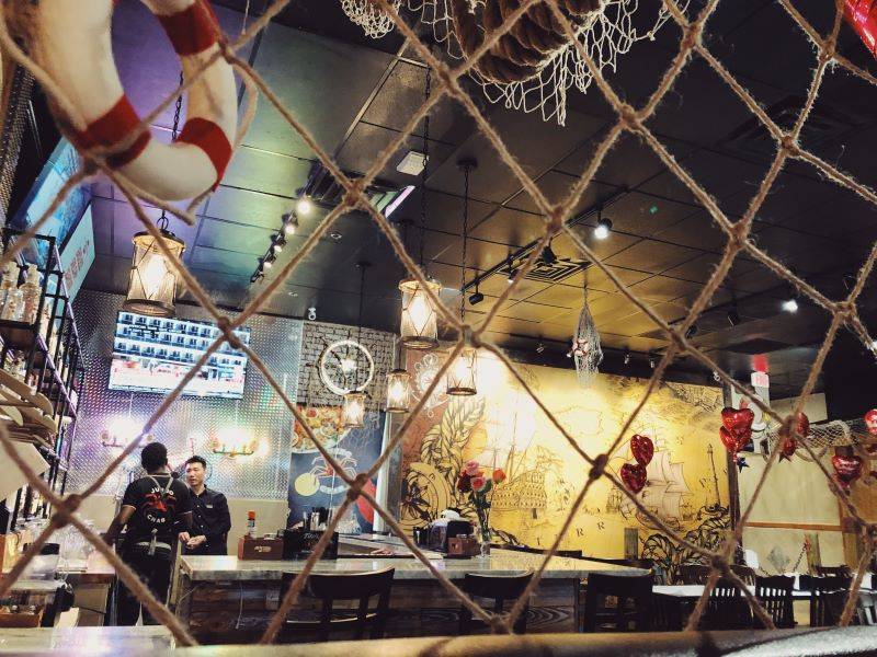 A fishing net spreads across the image of Jumbo Crab's dining room. There are several empty tables and a black ceiling. In the background, there is a white and red striped life preserver. Two servers talk in the background next to a bar. Photo by Remington Rock.