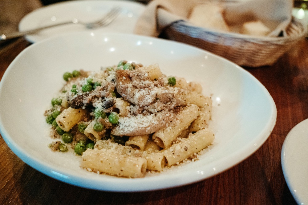 White plate of yellow pasta sits on top of a brown wooden table. There are green peas and grown mushrooms and sausage in the dish. There's a light brown basket in the back right of the photo. Photo by Anna Longworth.