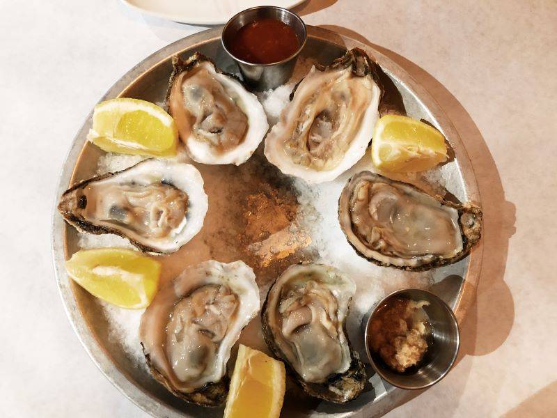 Six raw oysters arranged in a circle sit in an ice filled platter. Between each oyster is a lemon wedge. There are small sides of horseradish and cocktail sauce. Photo by Remington Rock.