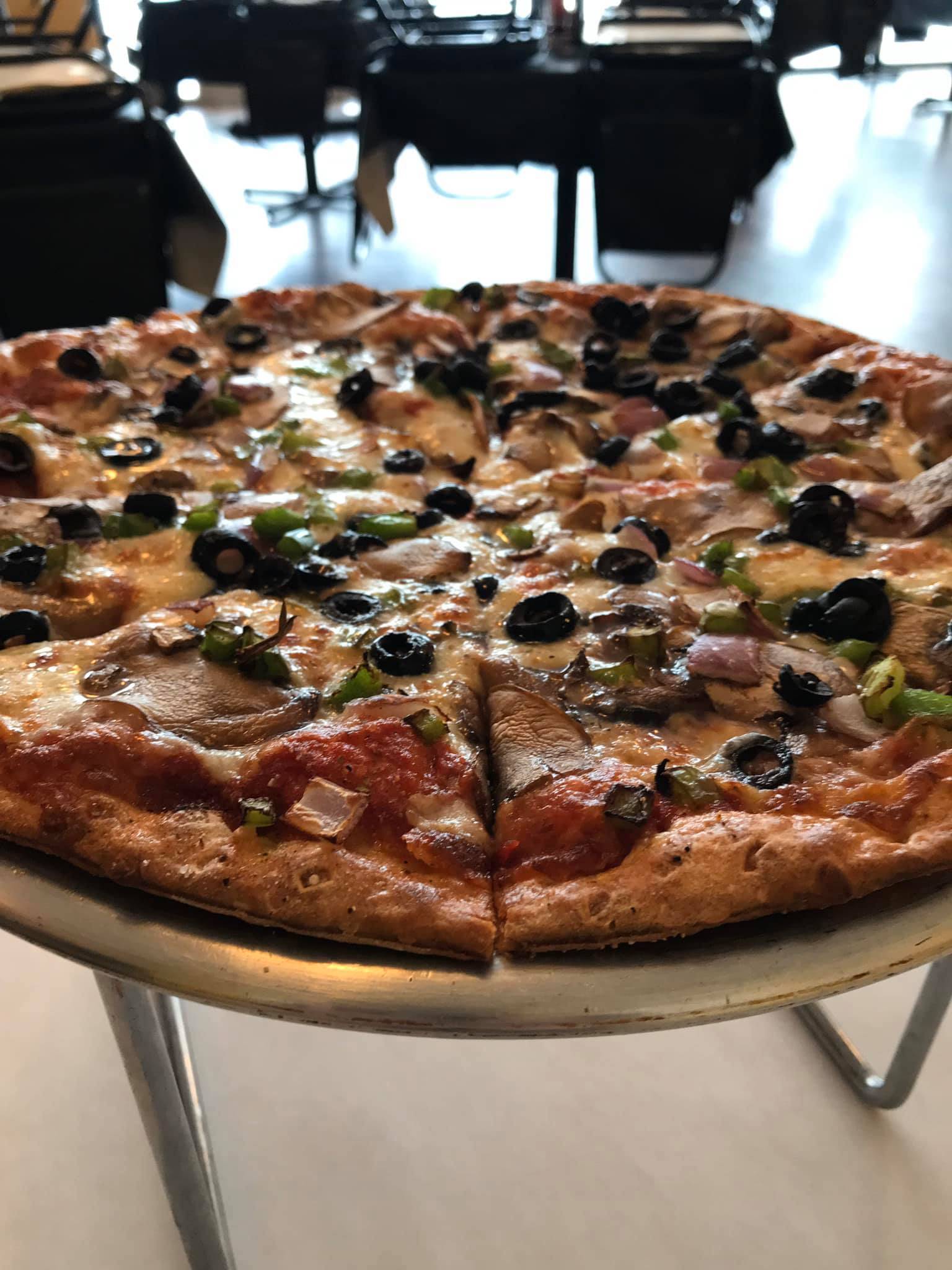 A large pizza pie with onions, green peppers, and olives is on a metal plate raised above a table. Photo from Po Boys Facebook page.