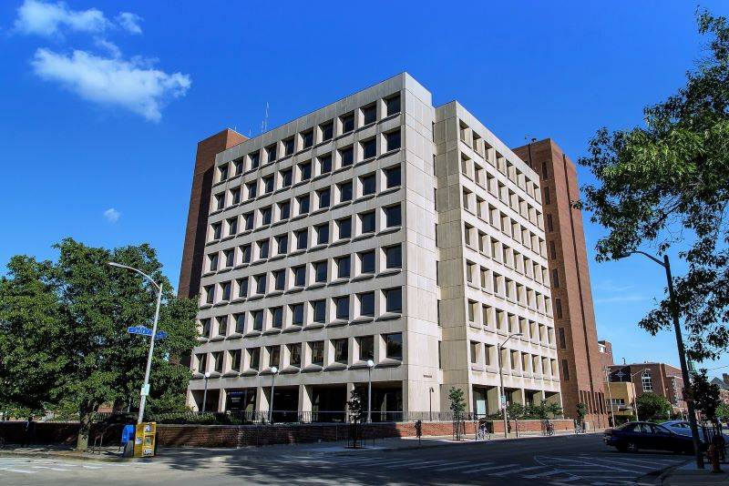 A light gray building with a grid of square windows on each visible face. It has red brick towers on either side. In the background is blue sky, and there are a couple of leafy green trees near the building. Photo from U of I Department of Psychology Facebook page.