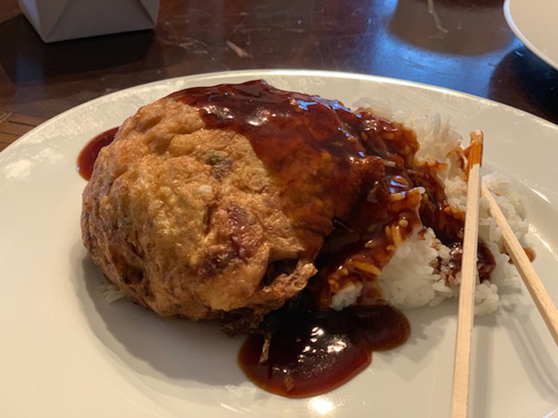 A crispy egg foo young sits on a cream colored plate with a dark brown sauce on top of part of the egg foo young. Touching and next to it, there is a small mound of white rice. Two wooden chopsticks are on the food. Photo by Seth Fein.