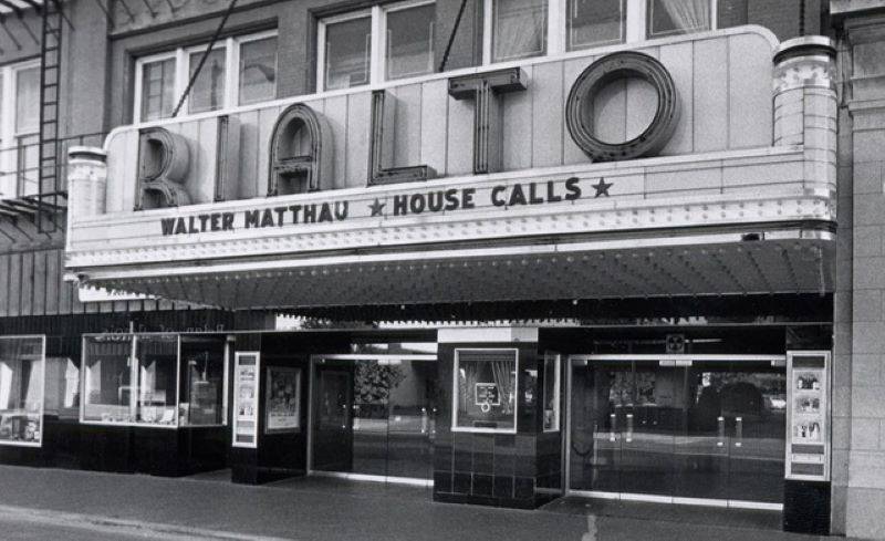 A black and white photo of the facade of a movie theater. The marquee has the word RIALTO in large letters. There are glass double doors and a box office window underneath.