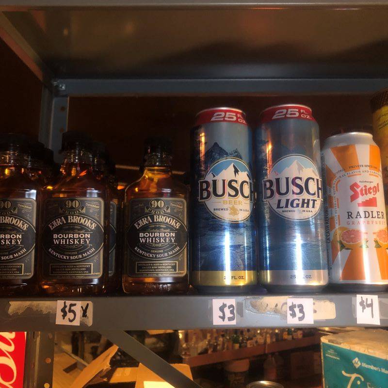 A metal shelf with bottles of bourbon and cans of Busch Light and Stiegl Radler. 