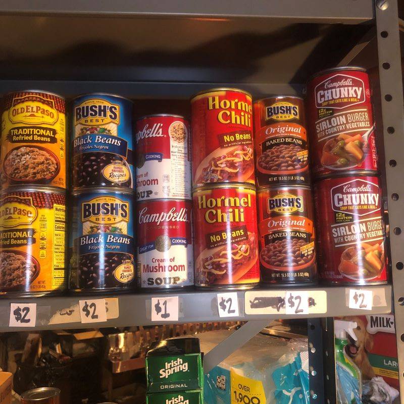 A metal shelf with cans of refried beans, black beans, cream of mushroom soup, Hormel chili, baked beans, and sirloin burger soup.