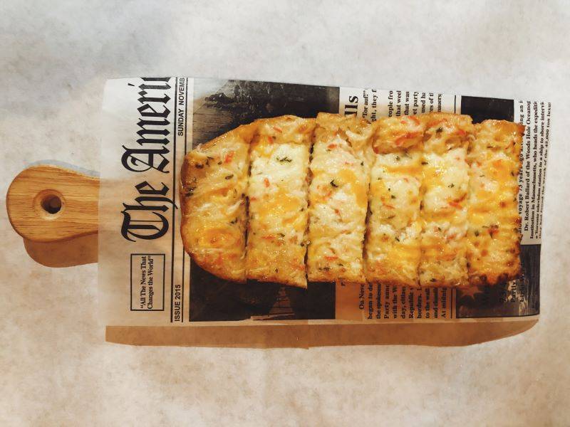 Six slices of cheesy seafood bread sit on a newspaper lined wooden board with a handle. Photo by Remington Rock.