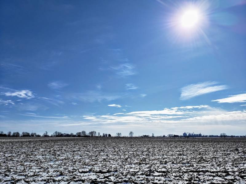 A vast field covered in a light layer of snow. There are trees dotting the horizon, and the sky is bright blue with wispy white clouds. Photo by Andrew Pritchard.