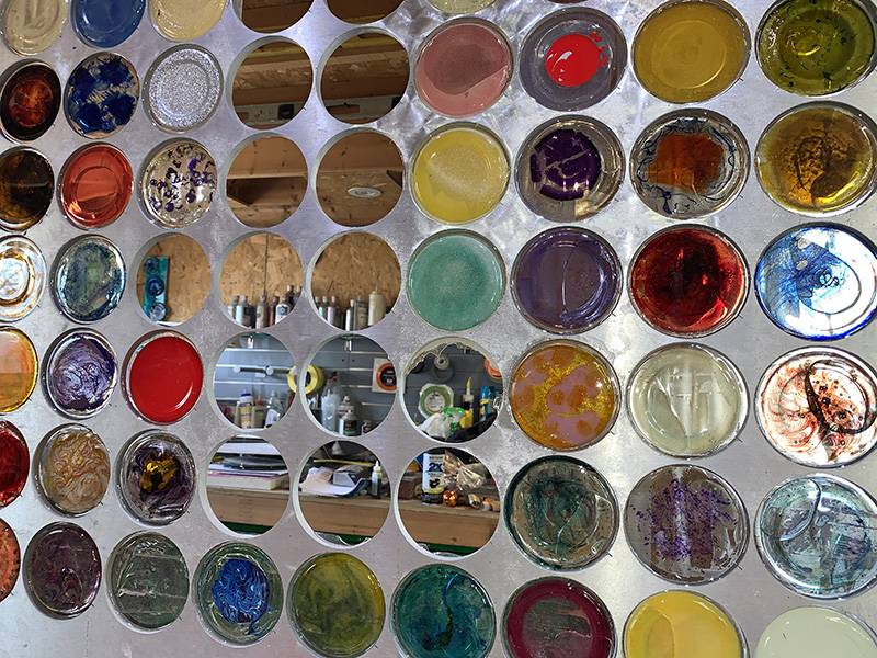 Image: Photo of current work in progress featuring a metal sheet with dozens of circles, most filled with multicolored combinations of resin and automative paint. Photo by Debra Domal