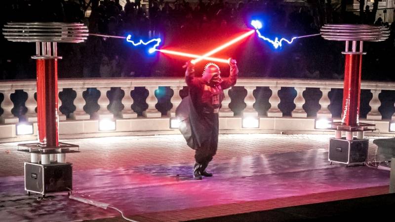 A person dressed as Darth Vadar holding two red glowing light sabers is standing between two large metal coils. There are electric currents coming out of each coil and reaching to the ends of the light sabers. Photo by Sam Logan.