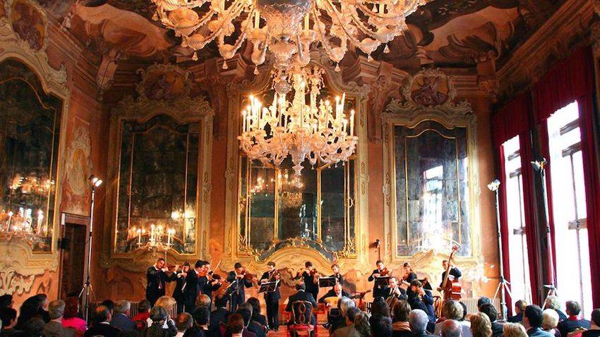 The Venice Baroque Orchestra plays in an ornately decorated room. A large chandelier hangs from the ceiling over a well-dressed audience. Light pours from the floor-to-celing windows, reflecting off large mirrors on the opposite walls.