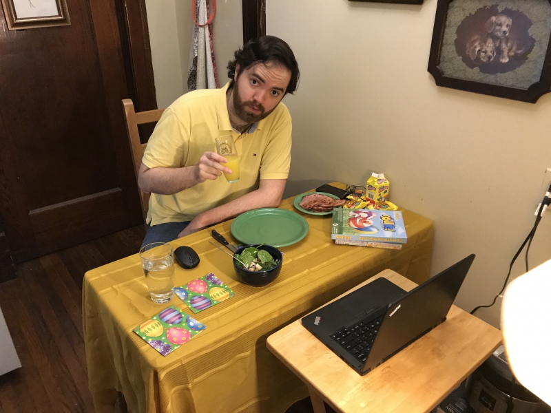 The author is sitting at a small table with a gold tablecloth, set for dinner. He has dark hair and a beard and is wearing a yellow shirt. A laptop is sitting open on a wooden tv tray in front of the table. Photo by Andrea Black.