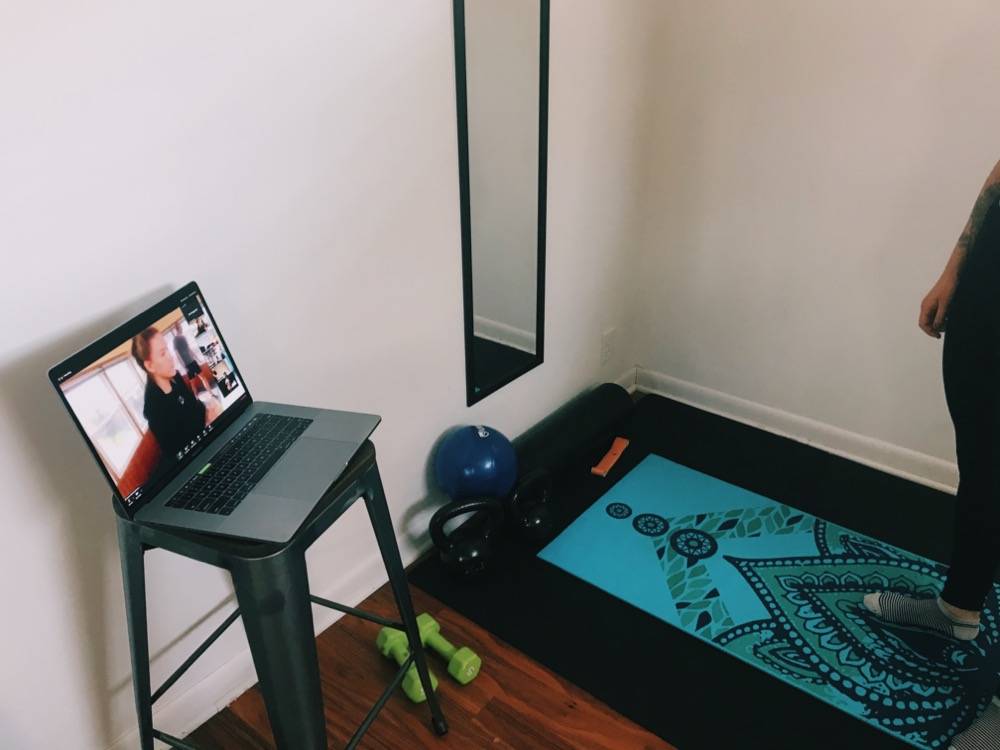 IMAGE: A laptop sits on top of a stool, there are a few pieces of workout equipment on the floor,  there's a black frame around a mirror on the white wall. There's a blue yoga mat on the ground. Photo by Patrick Singer.