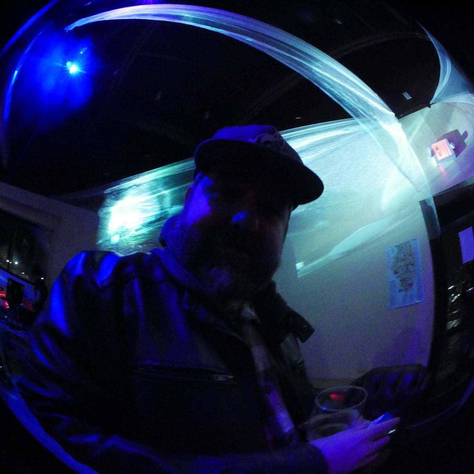 IMAGE: Dark image of a man wearing a black hat in a dark room with blue lights. There's an exit sign on the right side of the image. Photo by Matt Harsh.