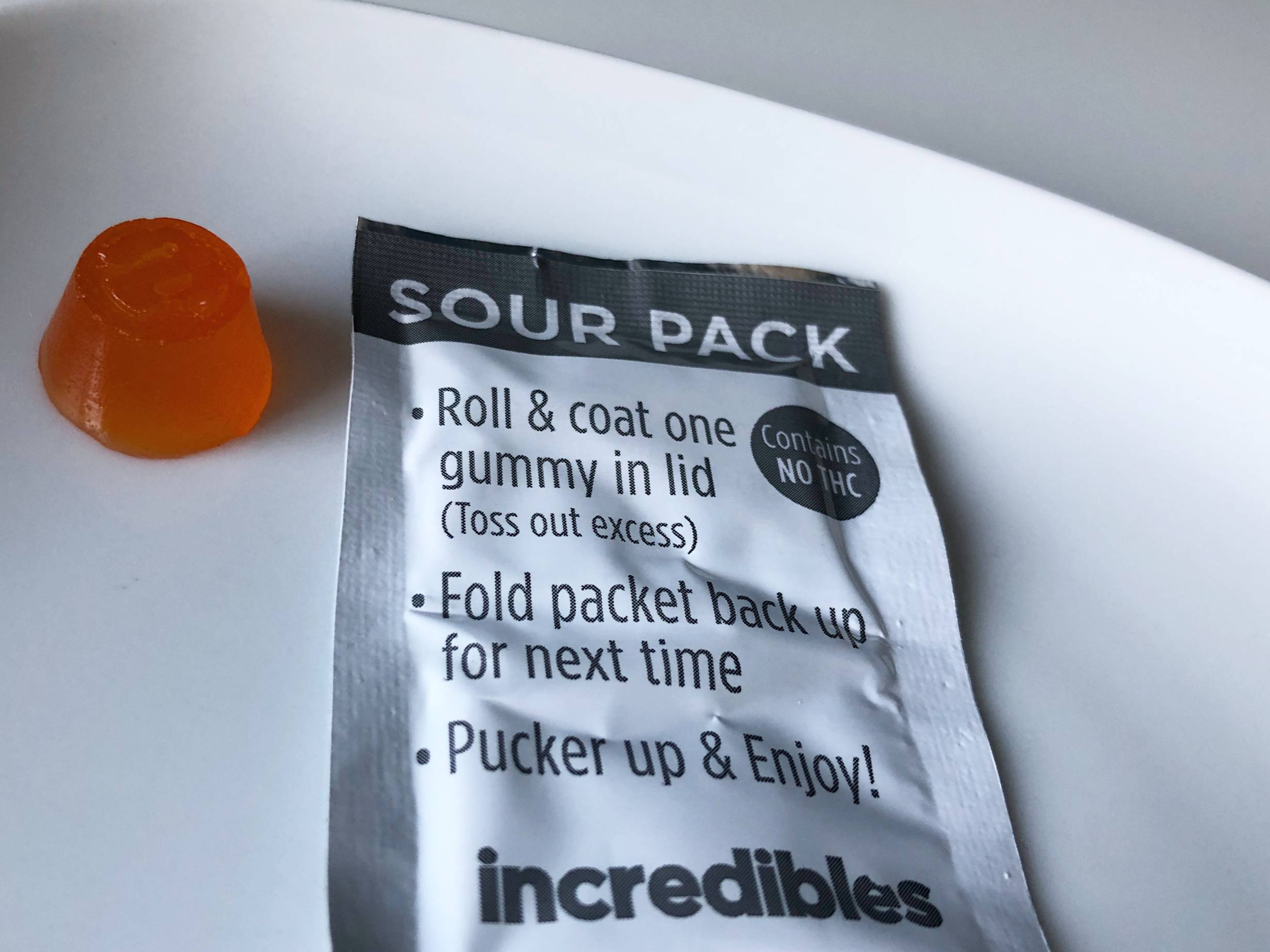 An orange cannabis-infused gummy sits on a white table next to a metal packet of sour-sugar with instructions for rolling the gummy in the packet. Photo by Alyssa Buckley.