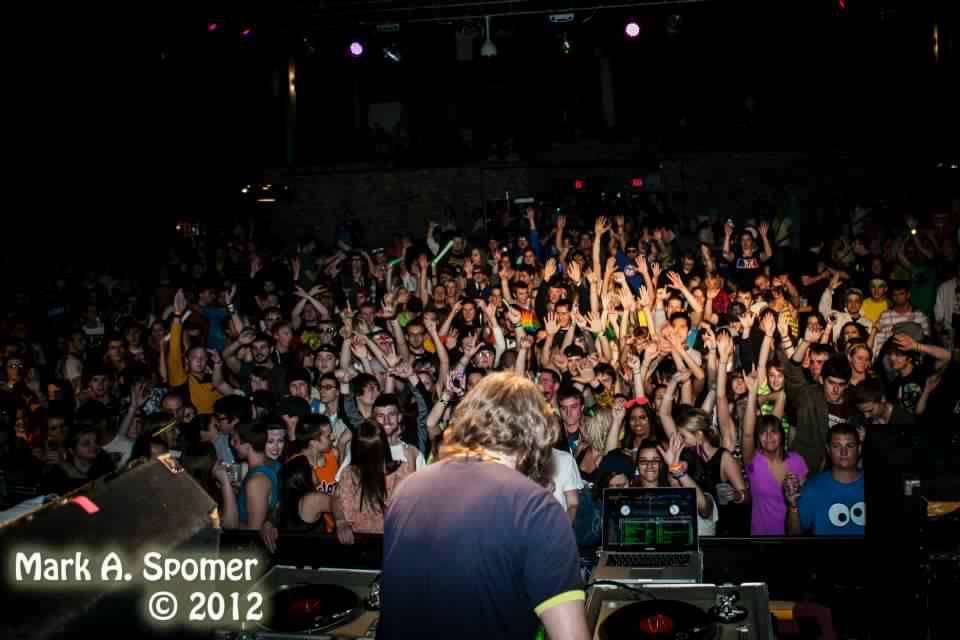 IMAGE: DJ with his back turned to the camera in the foreground, in the background is a crowd of people with their hands in the air. Photo by Mark A Spomer.