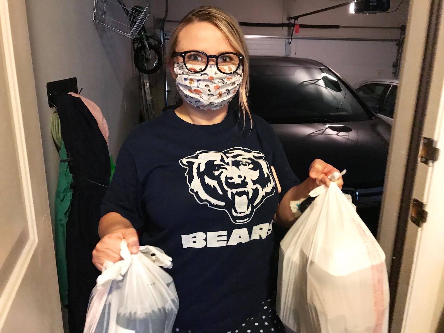 Alyssa Buckley wears a mask and holds two plastic bags of to-go orders. She stands in the threshold of garage and house with the door ajar revealing a dark garage. Photo by Alyssa Buckley.