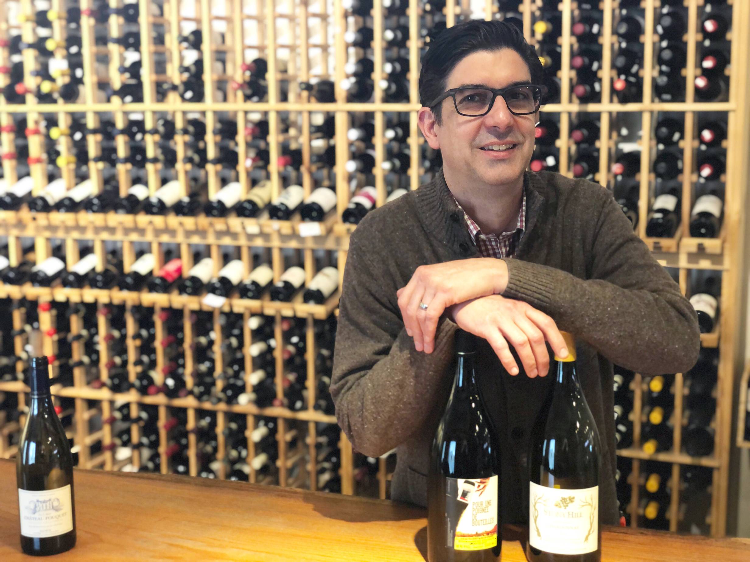 Todd Fusco stands over two bottles of wine on a wooden bar. He is wearing glasses and leaning both hands on the bottles. Behind him are wooden shelves with hundreds of bottles of wine. Photo by Alyssa Buckley.