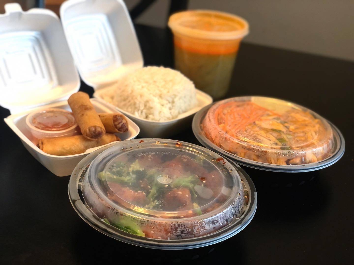 Two small styrofoam containers are open: eggrolls in one and white rice in the other. Two circle containers are closed with pad thai and sesame chicken. There is a tall, plastic cylindrical container holding yellow curry on a black table. Photo by Alyssa Buckley.
