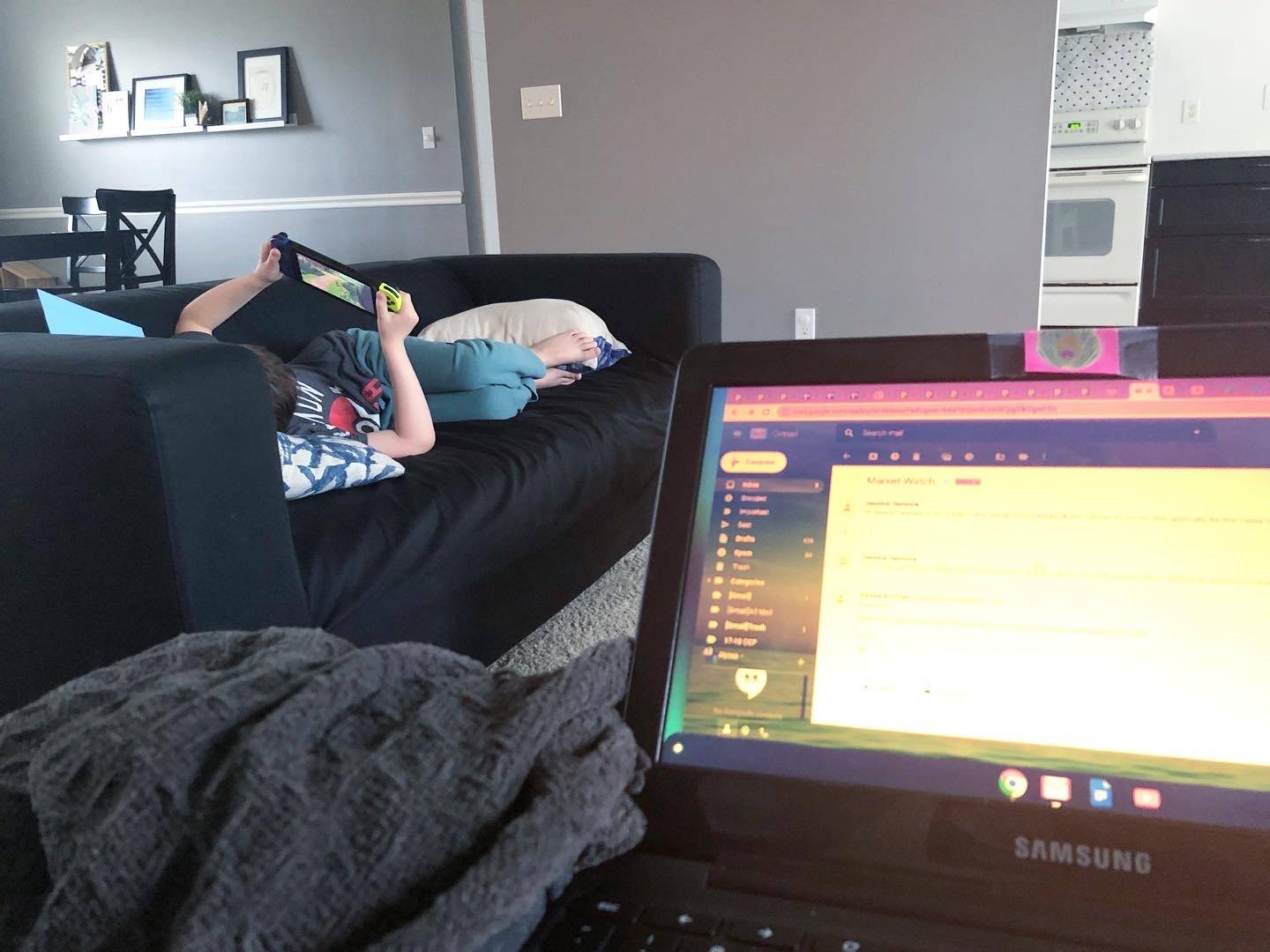 A child lays on a black couch with a Nintendo Switch. There is an open laptop with a screen too blurry to read in the lap of the photographer with a gray waffled blanket. Photo by Alyssa Buckley.