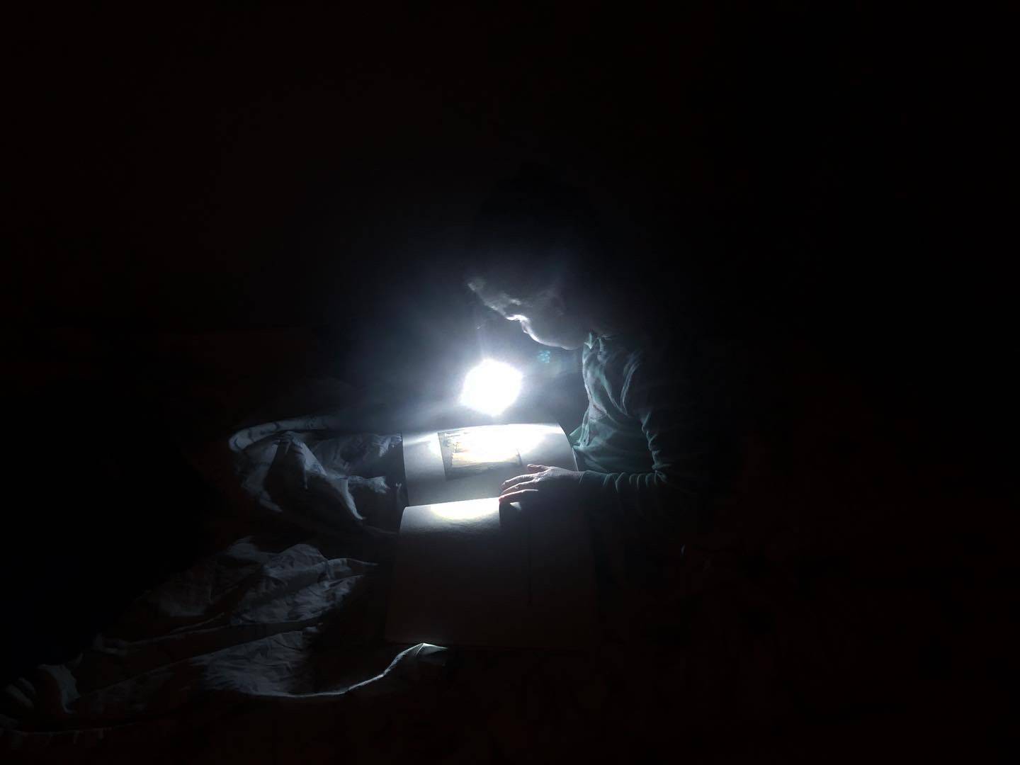 A dark room with a child seated in bed. The child is reading a book by the light of a flashlight. Photo by Alyssa Buckley.
