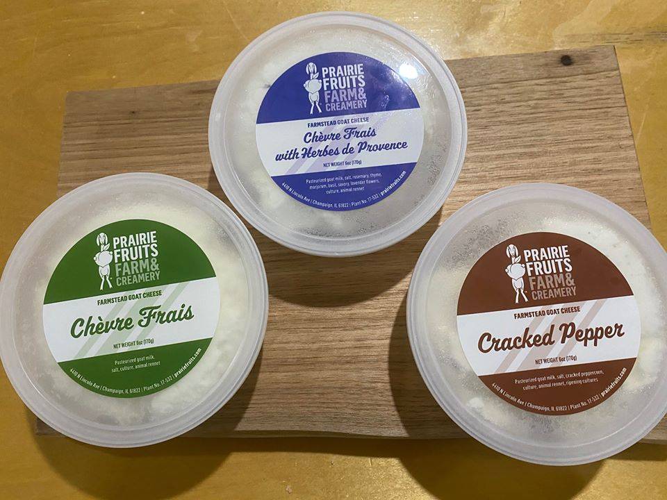 Three circular plastic containers of chevre cheese sit on a wooden cutting board. The label has a goat stirring a bowl next to the text of 
