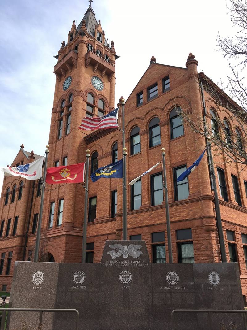 Photo of the present Courthouse from the northwest corner looking up to the clock tower.  Three stories tall, red sandstone and mottled brick, in a Romaneque style of architecture.  The Champaign County Veterans Memorial is in the foreground, with flags flying representing all the branches of the armed services.  Photo by Rick D. Williams.
