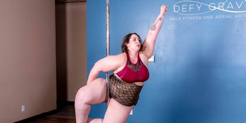 A woman is hanging on a metal pole, and one arm is outstretched with her hand in a fist. She is wearing a red sports bra style top and bronze mermaid pattern dance shorts. On the blue wall in the background it says Defy Gravity Pole Fitness and Aerial Arts in white lettering. Photo provided by Defy Gravity. 