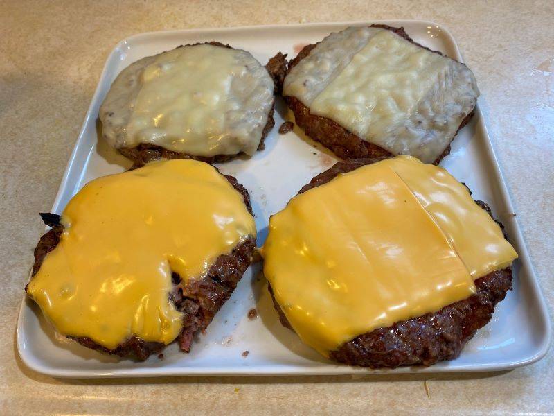 A white plate with four large hamburger patties. Two have melted Swiss cheese and two have melted American cheese. Photo by Julie McClure. 