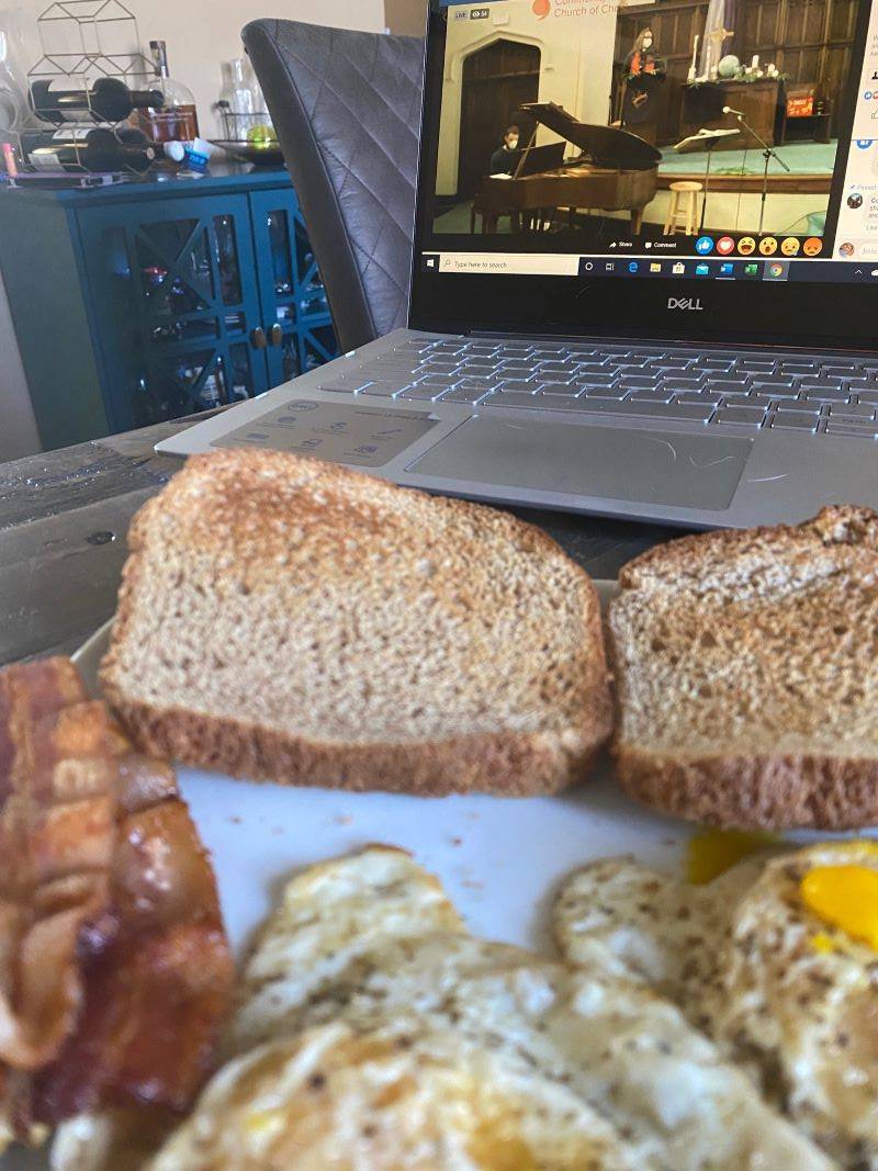 A partial view of an open laptop. On the screen is the altar of a church, with a woman pastor standing there wearing a mask. In the foreground is a white plate with eggs, bacon, and toast. Photo by Julie McClure.
