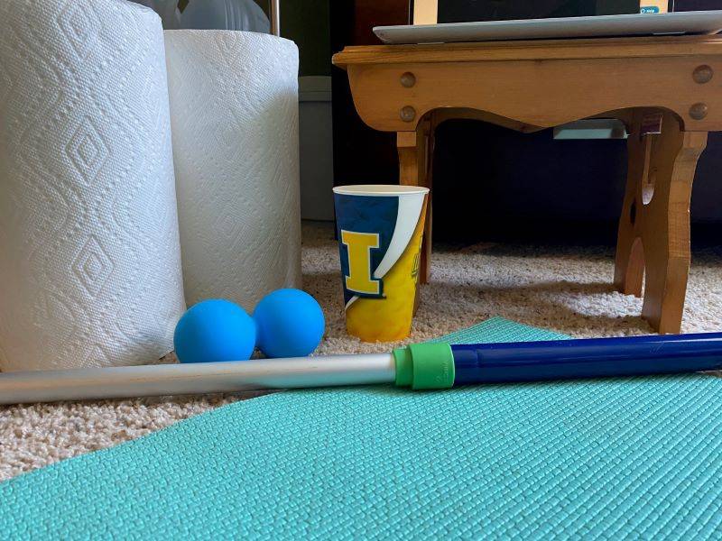 A collection of equipment on the floor: Two rolls of white paper towels, a blue peanut ball, a broom handle, a cup with an orange block 