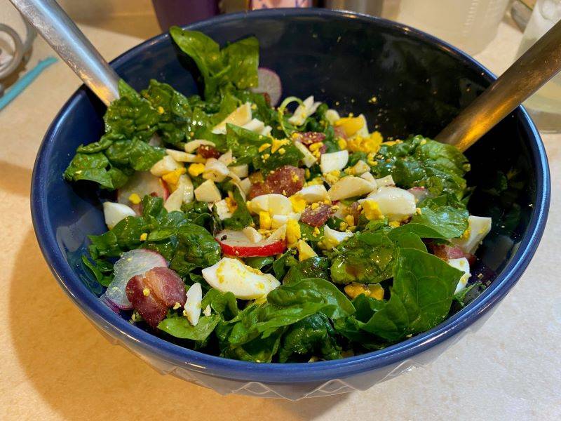 A blue bowl filled with salad. It has spinach, thinly sliced radishes, chopped hard boiled eggs, pieces of bacon. There are two silver salad utensils sticking out of the bowl. Photo by Julie McClure.