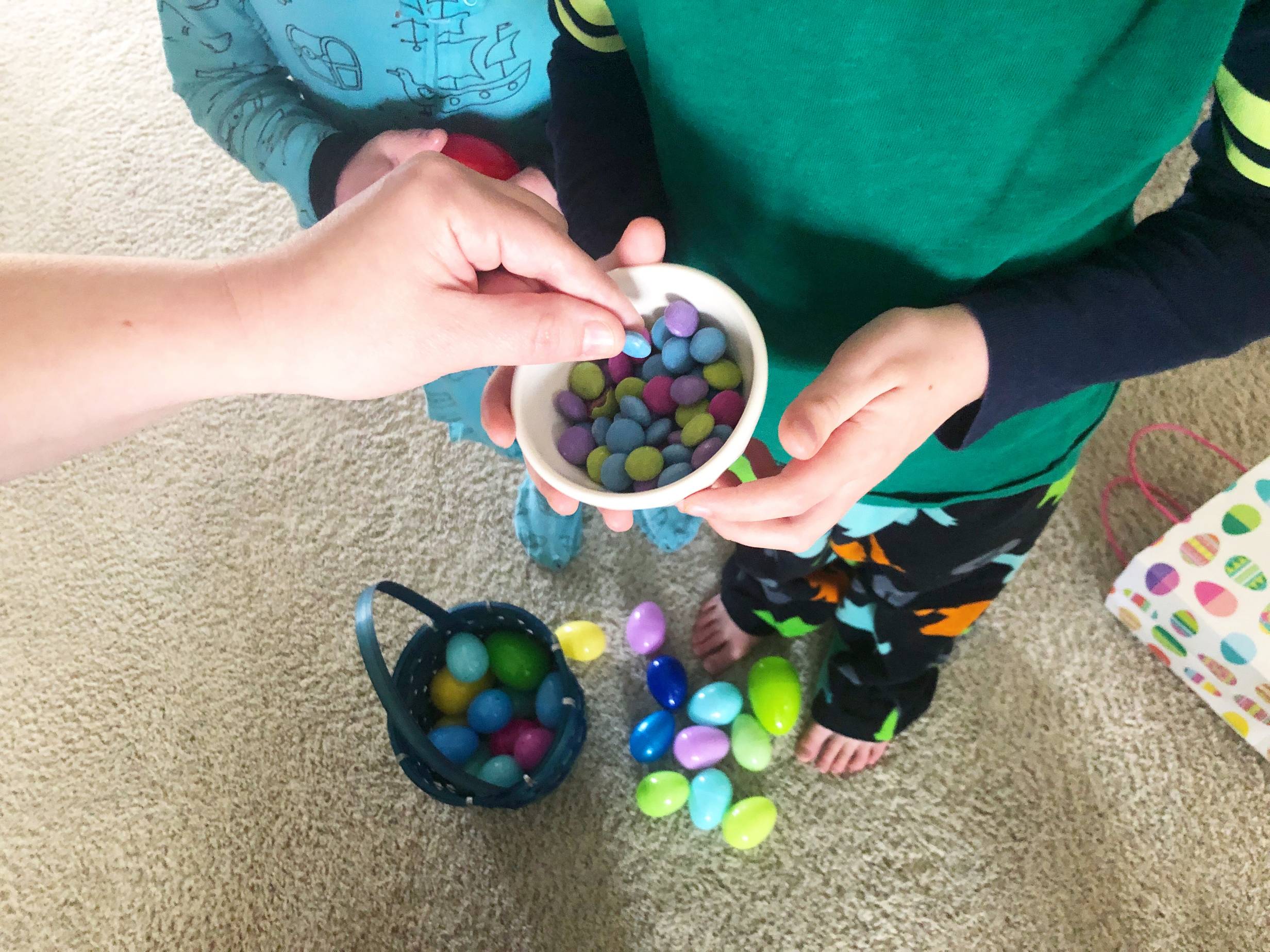 A bowl of multi-colored candy chocolates is held by a child in pajamas while an adult hand pinches a candy from the bowl. On the floor, there is a blue easter basket, many easter eggs, and a paper bag with easter eggs on it. Photo by Alyssa Buckley.